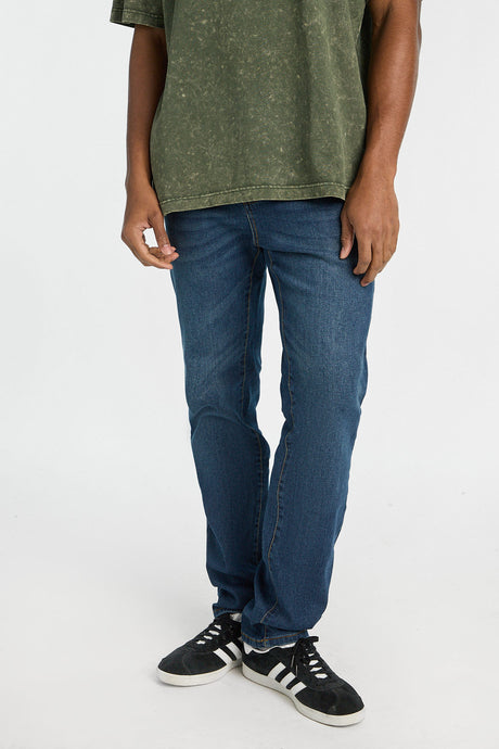 Male model in a green acid wash tee and Brodie Slim Straight jeans in blue with black sneakers.